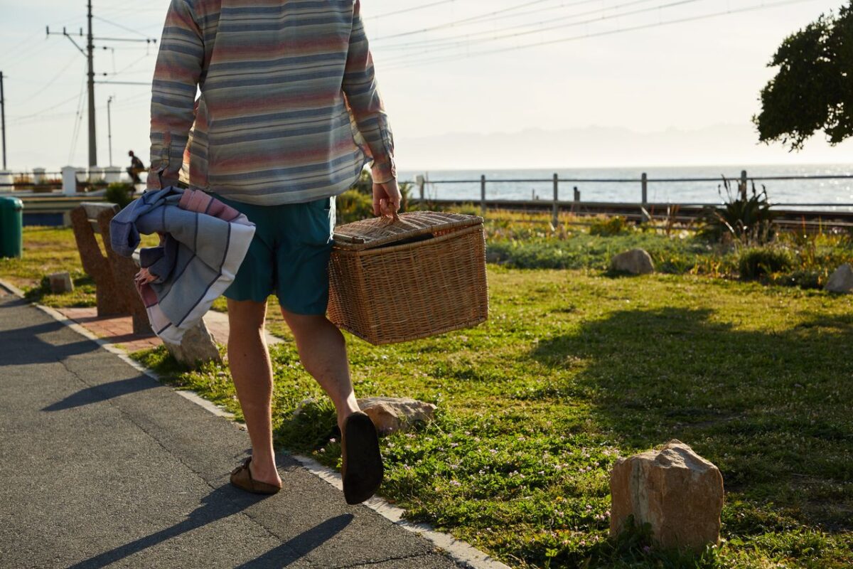 SB Déco Ambiance A person strolls on a sunny park path beside the ocean, carrying a picnic basket and blanket.