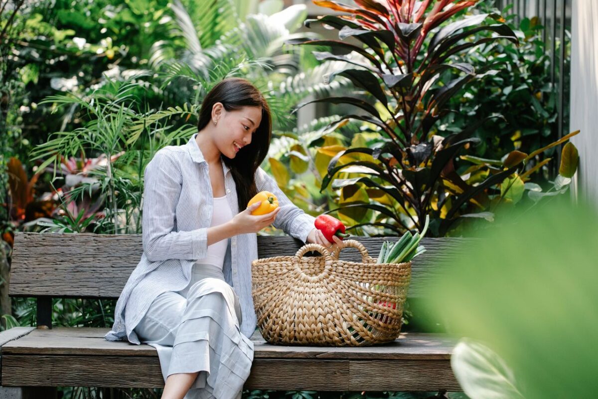 room-02 Content Asian female wearing casual outfit placing fresh yummy capsicums in wicker basket while sitting on wooden bench in verdant garden